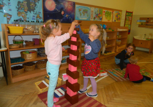 Dwie dziewczynki układaja wieżę z różowych sześciennych klocków i brązowych prostopadłoscianów (materiał montessoriański). W tle widać szafki z materiałem z wychowania kosmicznego oraz na ścianch plakaty z kontynentami i planetami układu słonecznego