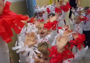 Dzieci stoją na korytarzu przed salą gimnastyczną- para za parą. Przedszkolaki ubrane są na biało- granatowo.Do bluzkek mają przymocowane kotyliony w barwach narodowych a wokół nadgarstków czerwone i białe opaski z frędzlami z bibuły.