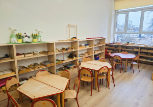 Klasa przedszkolna- pod scianą stoją otwarte szafki z pomocami montessoriańskimi. Po prawej stronie okno a pod nim stoją szafki również z pomocami. Na środku znajdują się stoliki z krzesełkami dostosowanymi do wzrostu dzieci