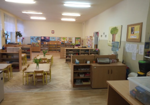Na zdjeciu pokazana jest klasa przedszkolna z szafkami na których znajdują się w koszykach pomoce rozwojowe. Po środku i po prawej stronie widać stoliki z wazonami, w których zanajdują sie tulipany.
