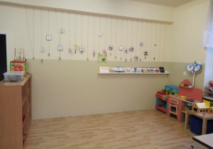 Klasa przedszkolna na której pokazana jest ściana z umieszczonymi na sznurkach zdjęciami dzieci. Pod nimi znajduje się półka z książkami. Po prawej stronie drewniana kuchenka dla dzieci, warsztat stolarski i gospodarstwo.