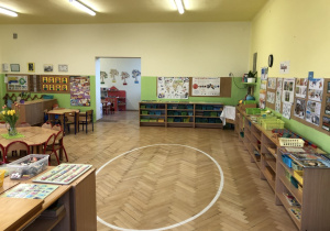 Klasa przedszkolna, w której znajduje sie biała namalowana linia w kształcie elipsy. Dookoła przy ścianach znajdują się szafki z półkami na których w koszykach znajdują się pomoce rozwojowe. Ściany do połowy wysokości pomalowane są na jasno-zielony kolor. W dali widać wejście do drugiego pomieszczenia.