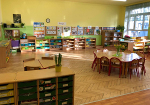 Klasa przedszkolna ze stolikami ustawionymi w sześciokąty. N tylnej ścianie znajdują się plakaty i zdjęcia dydaktyczne.