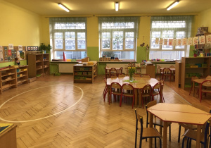 Klasa przedszkolna z szafkami i stolikami dla dzieci. W oddali widać trzy okna. Po prawej stronie na sznurku wystawa prac dzieci.