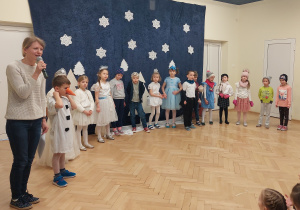 Dzieci przebrane za różne postacie stoją w pókolu, nauczycielka mówi przez mikrofon.