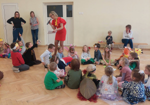 Nauczycielka mówi przez mikrofon do dzieci siedzących w kołach na podłodze.