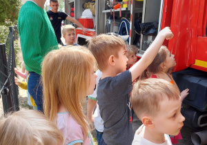 Dzieci stoją przed wozem strażackim i ogladają jego wyposażenie.
