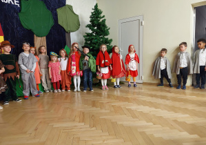 Grupa dzieci stojących w półkolu biorących udział w przedstawieniu Czerwony kapturek. Dzieci przebrane za gajowych, ptaki, Czerwone Kapturki, wiewiórki.