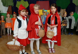 Trzy dziewczynki przebrane za Czerwone Kapturki. W tle dzieci biorące udział w przedstawieniu, przebrane za ptaki.