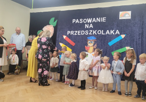 Pani Dyrektor pasuje nowe przedszkolaki dotykając ich ramienia dużym ołówkiem. Pani Wicedyrektor zakłada każdemu dziecku medal.