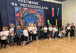 Dzieci stoją w półkręgu na tle kotary z dekoracją z okazji Dnia Przedszkolaka