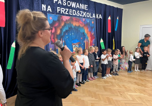 Pani Karolina mówi do dzieci prze mikrofon. Dzieci, które stoją w półkręgu klaszczą