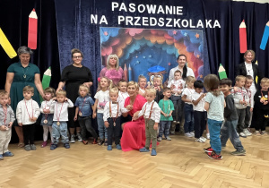 5 pań stoi przed kotarą z napisem Pasowanie na Przedszkolaka. Przed paniami stoją nowe dzieci .