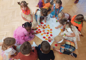 Dzieci siedzą przy dużym kartonie i wyklejają liśćmi sukienką pani Jesieni