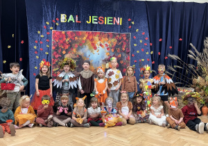 Na tle kotary z napisem Bal Jesieni stoją a przed nimi siedzą dzieci z grupy 3.