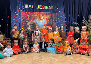 Na tle kotary z napisem Bal Jesieni stoją dzieci z grupy 4 odpowiednio przebrane do tematu balu