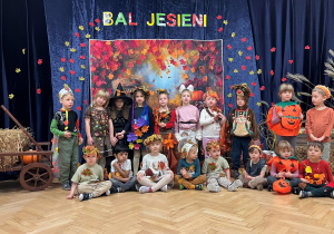Na tle kotary z napisem Bal Jesieni stoją dzieci z grupy 2 w strojach jesiennych