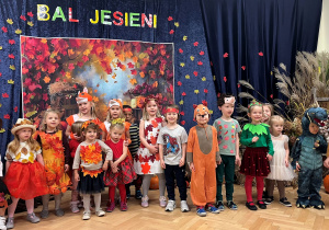 Na tle kotary z napisem Bal Jesieni stoją dzieci z grupy 5 przebrane odpowiednio do tematu balu