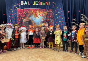 Na tle kotary z napisem Bal Jesieni stoją dzieci z grupy 7 przebrane w stroje o tematyce jesiennej