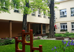 Budynek przedszkolny z ogrodem w którym znajdują się krzewy ozdobne, kwiaty i trawa.