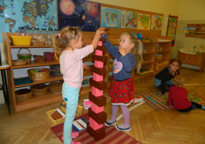 Dwie dziewczynki układaja wieżę z różowych sześciennych klocków i brązowych prostopadłoscianów (materiał montessoriański). W tle widać szafki z materiałem z wychowania kosmicznego oraz na ścianch plakaty z kontynentami i planetami układu słonecznego