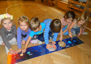 Sześcioro dzieci siedzi na drewnianej podłodze mając przed sobą układankę z puzzli przedstawiającą planety układu słonecznego.