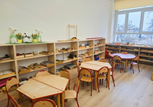 Klasa przedszkolna- pod scianą stoją otwarte szafki z pomocami montessoriańskimi. Po prawej stronie okno a pod nim stoją szafki również z pomocami. Na środku znajdują się stoliki z krzesełkami dostosowanymi do wzrostu dzieci