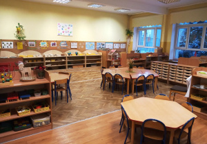 Klasa przedszkolna-trzy zestawy stolików ustawione w sześciokąt. Po lewej i prawej stronie znajdują się szafki z pomocami, na ścianie na tablicy korkowej znajdują się obrazki i plakaty.