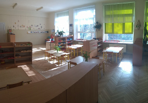 Klasa przedszkolna, w której znajdują się szafki z pomocami, stoliki dla dzieci. Okno zasłonięte jest przed słońcem jasno zieloną roletą.