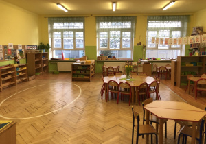 Klasa przedszkolna z szafkami i stolikami dla dzieci. W oddali widać trzy okna. Po prawej stronie na sznurku wystawa prac dzieci.