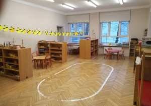Klasa przedszkolna na której środku znajduje się namalowana na podłodze biała linia w kształcie elipsy. Po bokach znajdują sie szafki otwarte z pomocami rozwojowymi.