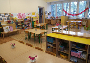 Klasa przedszkolna z sześcioma stolikami i krzesełkami. W oddali widać okna