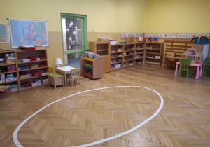 Klasa przedszkolna z namalowaną na podłodze białą linią w kształcie elipsy. Dookoła widać szafki z pomocami rozwojowymi.