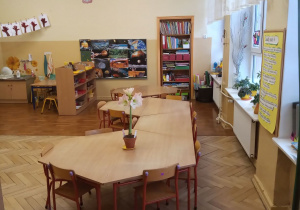 Klasa przedszkolna na środku stoją stoliki, dookoła szafki z pomocami. Na ścianie tablica z pracami dzieci.