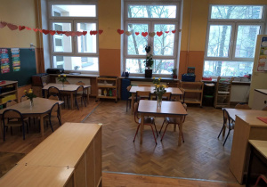 Klasa przedszkolna -z przodu znajdują się szafki z pomocami dla dzieci. Za nimi znajduje się pięć stolików z krzesełkami. W oddali znajdują sie trzy duże okna. Od okna do ściany powieszona jest girlanda z serduszek.
