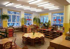 Klasa przedszkolna-na środku znajdują się stoliki ustawione w sześciokąt. Dookoła postawione są otwarte szafki z pomocami rozwojowymi. Na ścianach powieszone są paprotki a przy oknach balony.